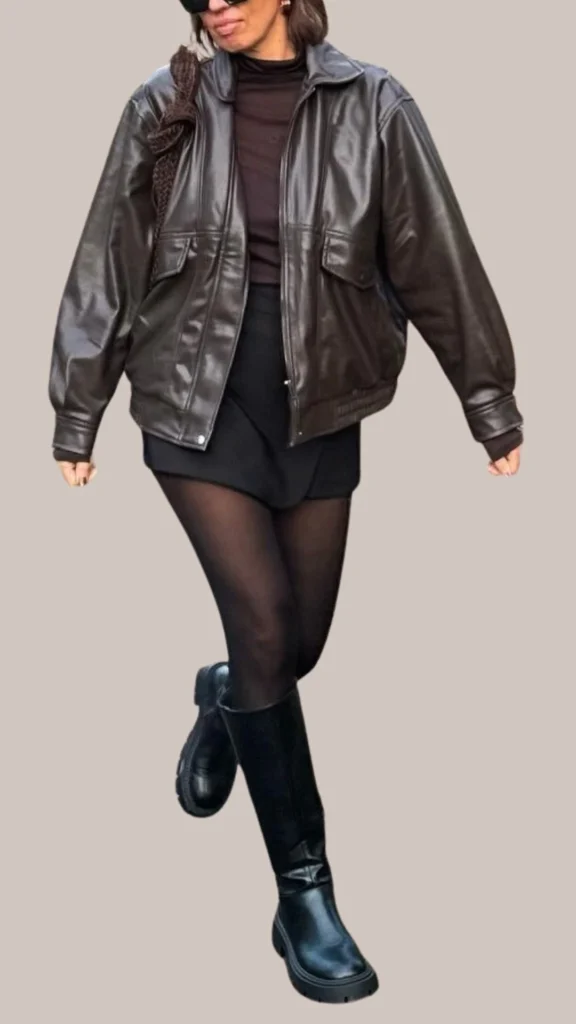 The outfit features a glossy, deep brown leather bomber jacket paired with a fitted, dark brown turtleneck and a sleek black mini skirt. She completes the look with sheer black tights and knee-high black leather boots, adding a touch of chic elegance, enhanced by minimalist silver hoop earrings and oversized black sunglasses.