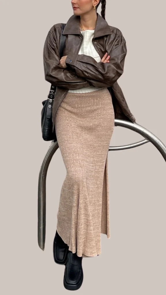 A rich, dark brown leather jacket with a structured collar is layered over a light cream ribbed sweater, providing a chic contrast. The outfit is completed with a long, light beige knit skirt that features subtle texture, and sleek black chunky boots for a modern edge. A small black crossbody bag adds a functional touch to this stylish ensemble.