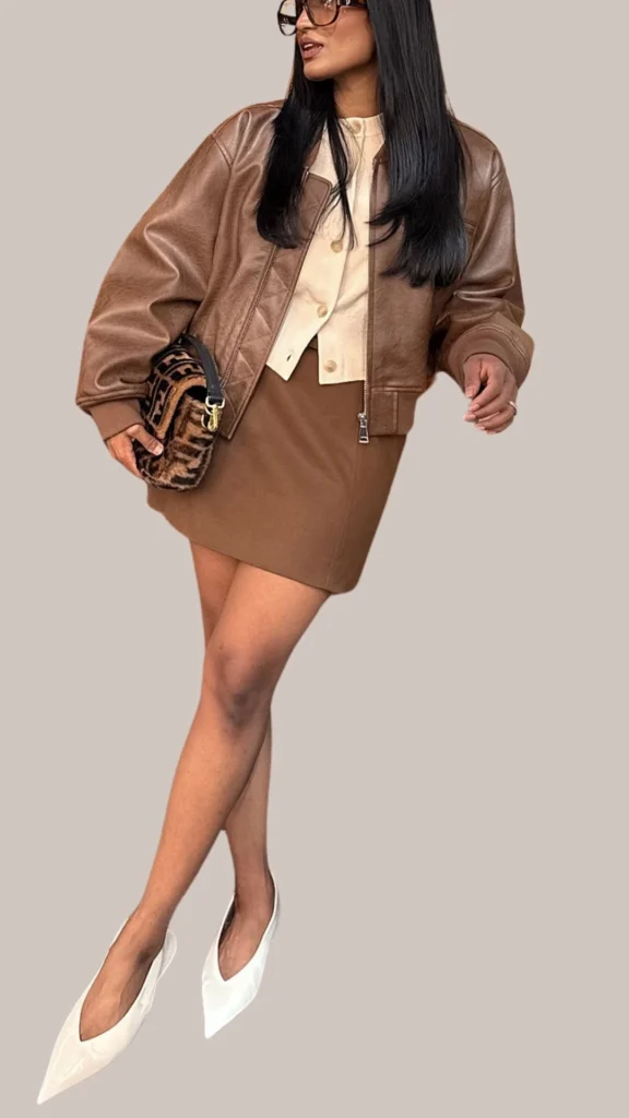 A chic outfit features a rich, dark brown leather jacket with a relaxed fit, paired with a lighter brown, form-fitting mini skirt. Underneath, a cream-colored cropped top with buttons adds a soft contrast, while sleek white pointed-toe heels complete the look, alongside a textured brown bag for added elegance. This ensemble combines various shades of brown with subtle textures for a polished and sophisticated appearance.
