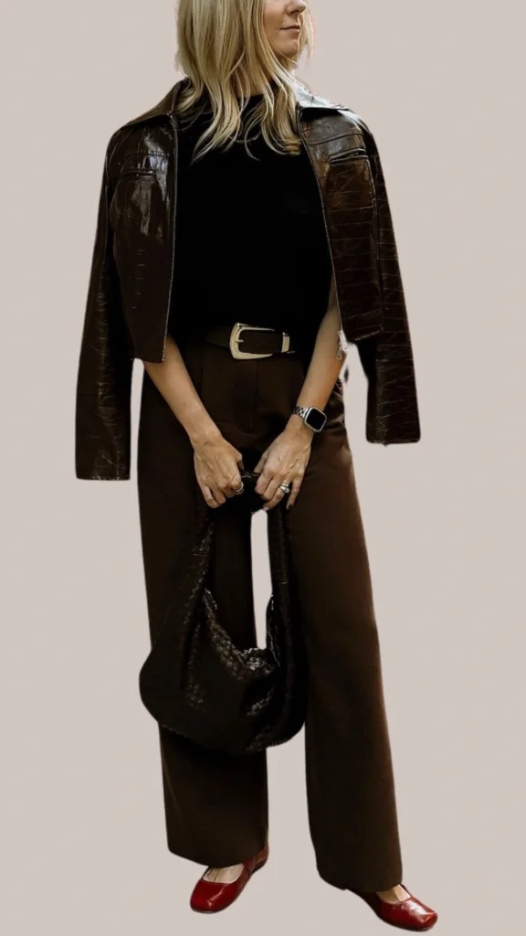 A chic brown leather jacket with a glossy finish is layered over a fitted black top, paired with high-waisted, wide-legged brown trousers that have a rich, warm tone. Complementing the outfit are deep red ballet flats and a textured black handbag, adding a touch of elegance and contrast. A silver belt and a watch complete the look, enhancing its stylish sophistication.