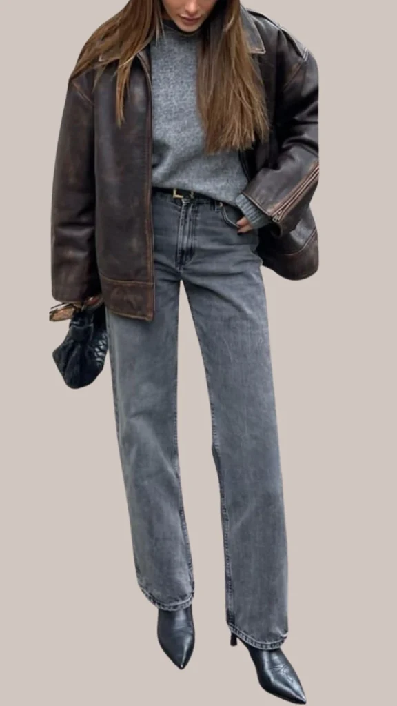 A dark brown leather jacket complements a soft gray sweater, exuding a chic layered look. The outfit is paired with high-waisted, slightly loose gray jeans and pointed black ankle boots, creating a stylish contrast of textures and colors. A small black handbag adds a refined touch, enhancing the overall elegant aesthetic.