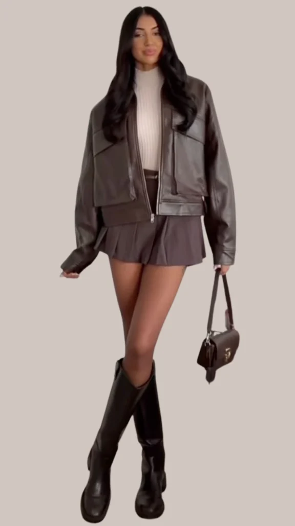 A chic brown leather cropped jacket is layered over a fitted cream ribbed turtleneck, paired with a flared gray mini skirt that adds a touch of contrast. The outfit is complemented by glossy black knee-high boots and a sleek brown handbag, enhancing the overall sophisticated look. Textures vary from the smooth leather to the soft knit and the structured skirt, creating a balanced ensemble.