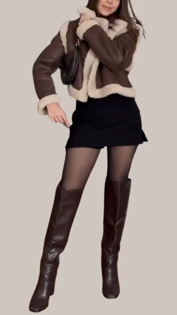 The outfit features a chic brown leather jacket with a soft, cream shearling collar and cuffs, paired with a sleek black mini skirt. The look is completed with thigh-high brown leather boots that add a touch of sophistication and texture contrast. A small black handbag enhances the ensemble, offering an elegant finishing touch.