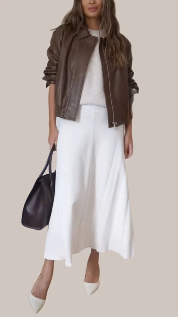 A rich, dark brown leather jacket with a smooth texture is layered over a soft, light gray sweater, paired with a flowing white midi skirt that has a slight sheen. Complementing the outfit are elegant white pointed-toe heels and a structured black handbag, creating a chic and sophisticated look. The combination of textures and contrasting colors enhances the outfit's polished appeal.
