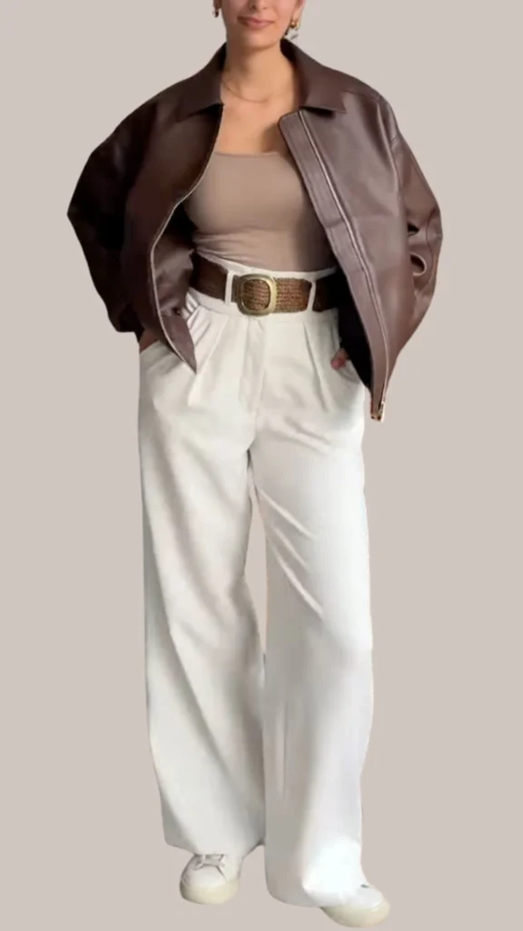 A stylish brown leather jacket with a soft, slightly distressed texture is paired with a fitted taupe tank top and high-waisted, wide-leg white trousers. The outfit features a woven tan belt that adds a polished touch, complemented by white sneakers for a chic yet casual look. The combination of rich brown tones with light neutrals creates a balanced and sophisticated aesthetic.