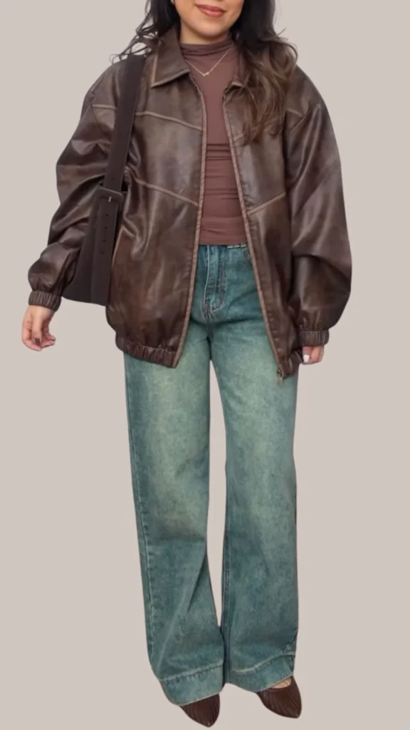 The outfit features a rich, dark brown leather jacket with a relaxed fit and subtle sheen, paired with a fitted, medium brown long-sleeve top underneath. The bottom half showcases light blue wide-leg jeans with a faded texture, creating a chic contrast, and the look is completed with stylish brown pointed-toe heels. A simple gold necklace adds a touch of elegance to the ensemble.