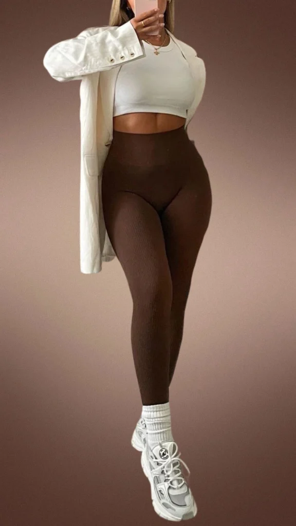 The outfit features high-waisted, ribbed brown leggings made from a cozy, stretchy fabric, paired with a cropped white top. A light-colored oversized blazer adds sophistication, while chunky white sneakers with contrasting details and white crew socks complete the look, making it both stylish and comfortable for winter.