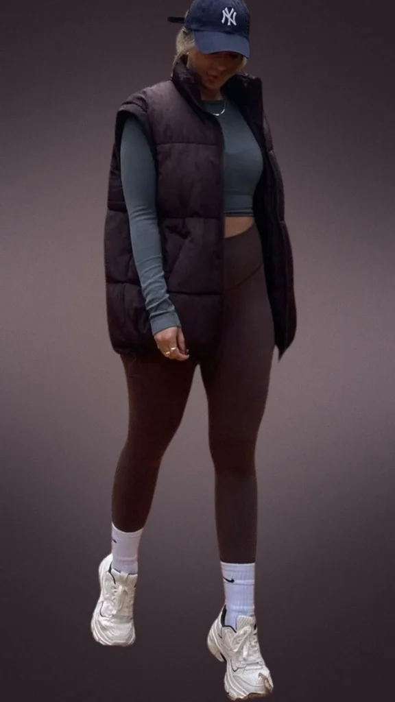 The outfit features a pair of snug brown leggings, complemented by a cozy black padded vest and a fitted teal long-sleeve top, ideal for winter wear. Accessories include a navy baseball cap and white ankle socks paired with stylish white sneakers, enhancing the overall comfort and casual look. The combination of soft fabrics and layered textures creates a warm and inviting appearance, perfect for cooler months.