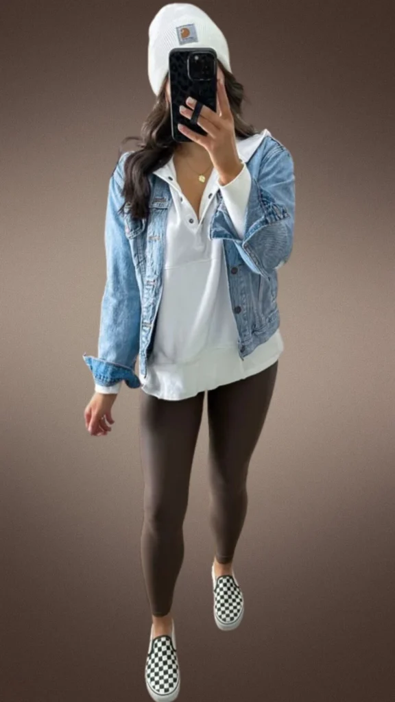 A cozy winter outfit featuring dark brown leggings made from a soft, stretchy fabric, paired with a white long-sleeve top layered under a light blue denim jacket. The look is complemented by a white knit beanie and black-and-white checkered slip-on shoes, creating a chic yet comfortable ensemble perfect for cooler months.