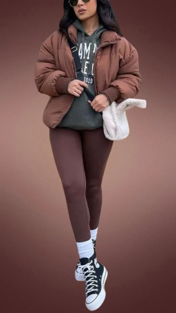 A cozy brown leggings outfit features ribbed, warm brown leggings paired with a dark green hoodie and a matching puffy brown jacket for insulation. The look is completed with high-top black sneakers and white crew socks, while a soft cream-colored bag adds a touch of texture, perfect for winter layering.