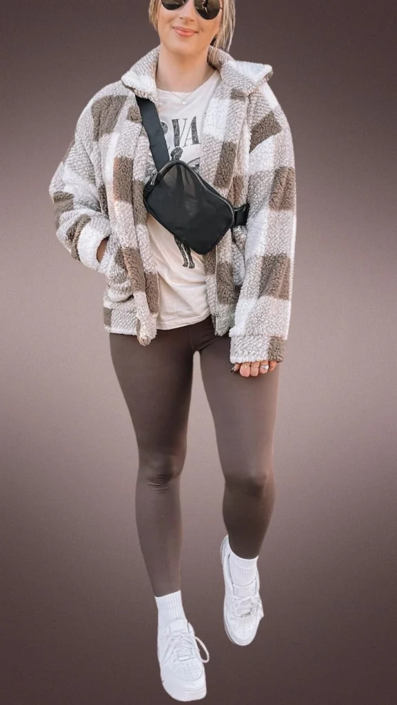 A cozy winter outfit featuring soft, plush brown striped leggings paired with a fluffy beige and brown plaid zip-up jacket. The look is completed with a white graphic t-shirt underneath, white ankle socks, and white sneakers, along with a sleek black crossbody bag for added practicality.