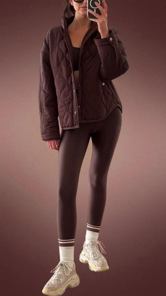 The outfit features snug, brown leggings made from a soft, stretchy fabric, paired with a cozy, quilted, aubergine puffer jacket that adds warmth. Complementing the look are chunky sneakers in light gray with yellow accents and white socks featuring maroon stripes, creating a stylish yet comfortable ensemble suitable for winter. The combination of textures and shades enhances the outfit's cozy appeal.