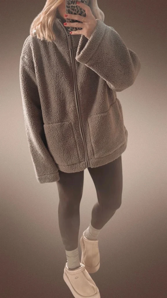 A cozy winter outfit featuring brown fleece leggings paired with a soft, oversized beige zip-up teddy coat, providing warmth and comfort. The look is completed with beige slippers and light grey socks, adding to the snug aesthetic ideal for cooler months. The textures of the fleece and leggings enhance the overall inviting feel of the outfit.