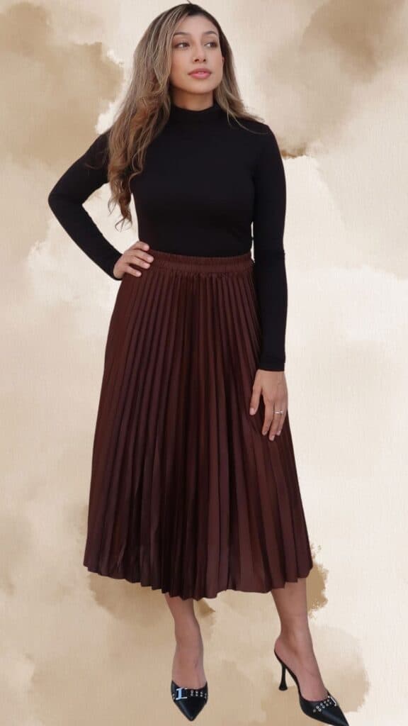 A fitted black turtleneck tops a chic, pleated brown midi skirt that flows elegantly with movement, showcasing a rich chestnut hue. The outfit is paired with stylish black stiletto heels featuring a decorative buckle, enhancing the classy vibe. A delicate silver ring adds a subtle touch of sophistication.