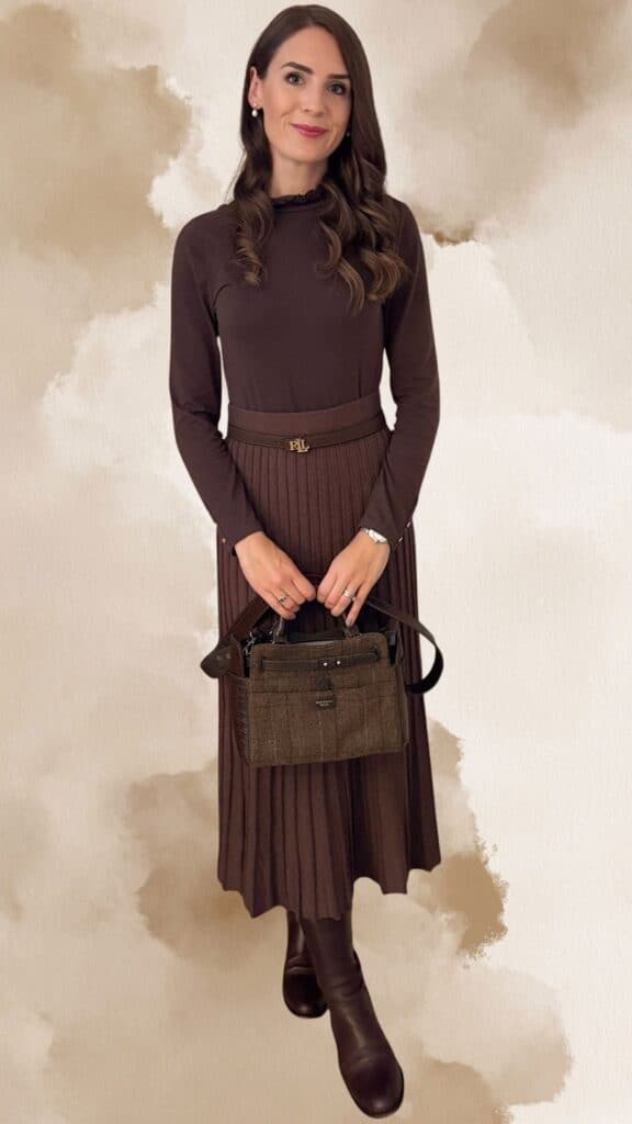 A fitted brown long-sleeve top pairs elegantly with a pleated brown midi skirt, showcasing a rich, deep chocolate hue. The outfit is accessorized with a matching brown belt featuring a stylish emblem, a textured brown handbag, and knee-high brown leather boots, creating a chic and sophisticated look. Subtle pearl earrings complement the ensemble, adding a touch of elegance.