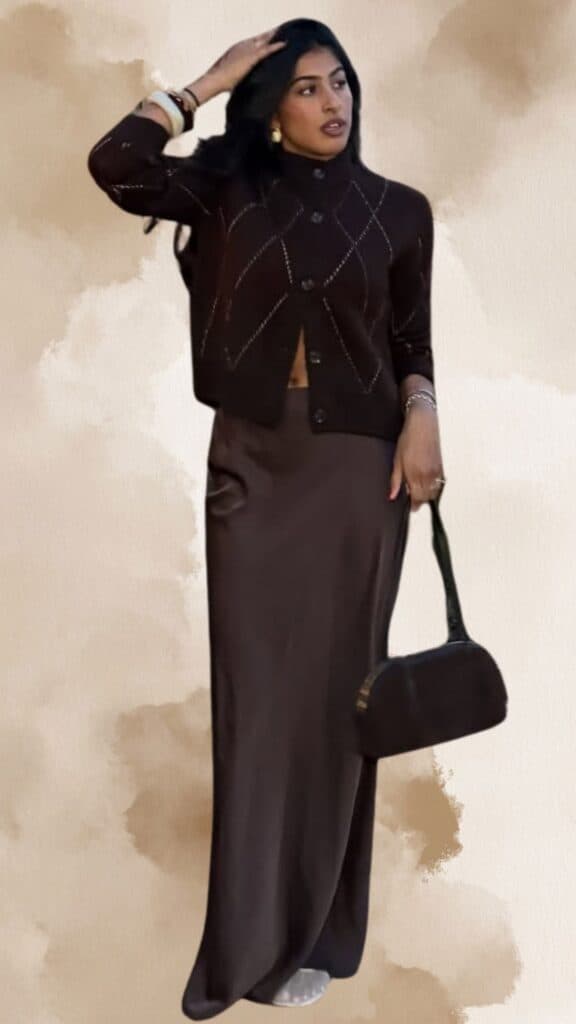 The outfit features a fitted, deep brown cropped cardigan with a diamond pattern and subtle texture, paired with a flowing, ankle-length brown satin skirt that exudes elegance. Accessories include stacked bangles and large hoop earrings, complemented by a chic, structured black handbag that adds a modern touch. This ensemble strikes a balance between comfort and sophistication, ideal for a classy look.