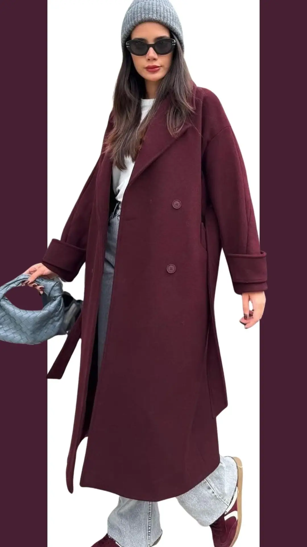The outfit features a long, double-breasted burgundy coat made from a soft, textured fabric, with wide sleeves and a relaxed fit. Underneath, it is paired with a simple white top and light-washed, loosely fitted jeans, complemented by laced burgundy sneakers. A cozy gray beanie and chic sunglasses enhance the modern, stylish look.