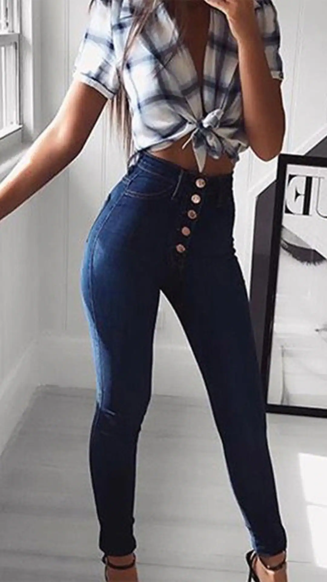 The outfit features high-waisted, form-fitting denim jeans with a deep indigo wash, enhanced by a unique front button closure and a sleek silhouette that extends to the ankle. Paired with a cropped, short-sleeve plaid shirt in white and light blue, this look is casual yet polished, perfect for a laid-back outing or a relaxed daytime event.
