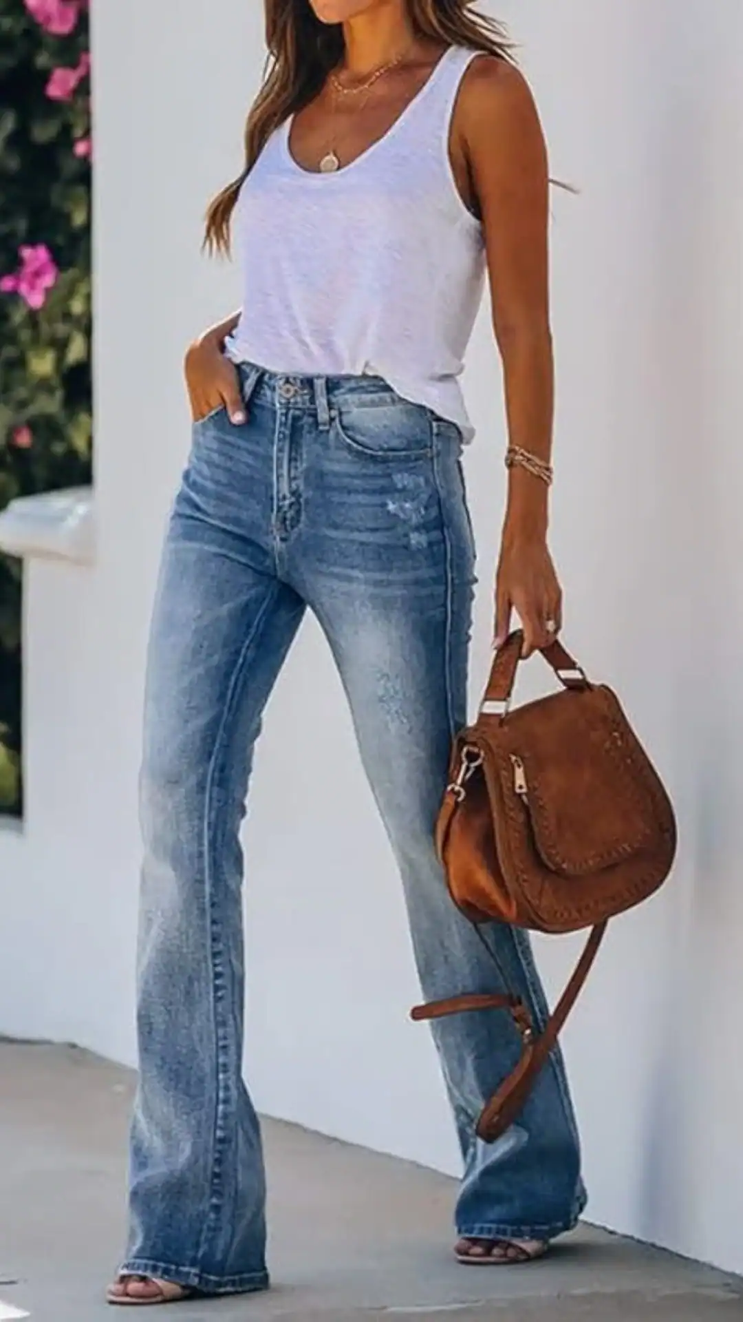 A woman showcases a casual summer look featuring high-waisted, flared light-wash jeans crafted from soft denim, complemented by a relaxed white tank top. The outfit exudes a laid-back vibe, perfect for a casual outing, accentuated by a chic brown leather crossbody bag and a minimalistic layered necklace.