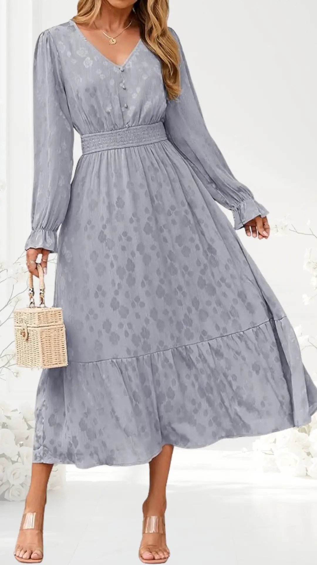 A long, flowing dress in a soft gray hue features a subtle floral pattern, long sleeves with gathered cuffs, and a stylish V-neckline with decorative buttons. The bodice is cinched with a smocked waistband, allowing for a flattering silhouette that flares out to a layered skirt. The outfit is complemented by a woven light-colored handbag and transparent heels, enhancing its elegant yet casual appeal.
