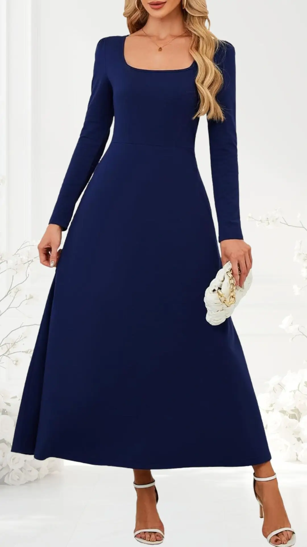 Long-sleeved navy dress features a square neckline and an A-line silhouette that flows elegantly to the floor. The model holds a white clutch and wears strappy, open-toed heels. The dress is complemented by a soft, minimal backdrop of floral accents.