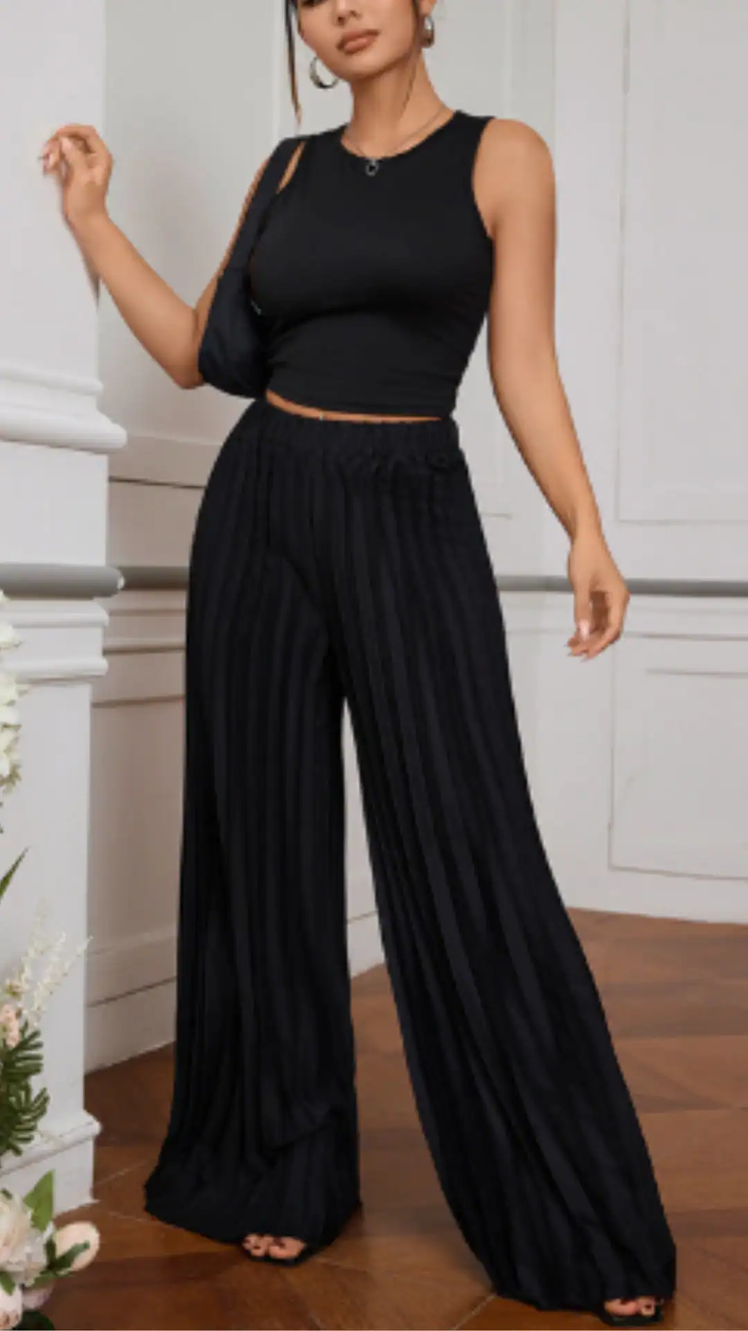 The outfit features high-waisted, wide-leg black pants made of a lightweight, textured fabric with vertical pleats that add movement. Paired with a fitted black crop top, this ensemble offers a chic and minimalist vibe, ideal for casual outings or evening events.
