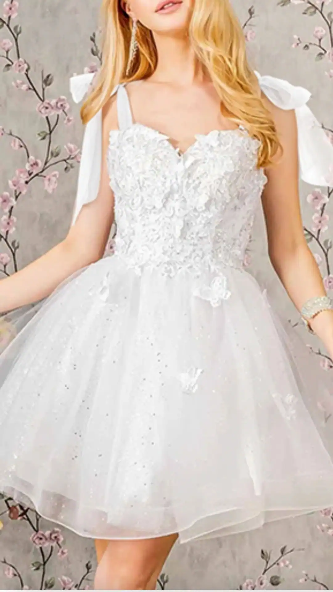 This chic white homecoming dress features a fitted, intricately beaded lace bodice and a voluminous tulle skirt that exudes a fairytale vibe, complemented by delicate butterfly appliqués. The dress is adorned with elegant, soft fabric shoulder ties, making it perfect for a festive occasion like homecoming or a school dance, appealing to teenage girls looking for a trendy yet romantic look.