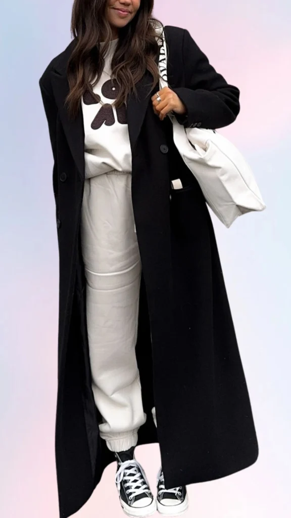 The outfit features a warm black oversized coat layered over a cozy white sweatshirt with a playful design, paired with soft, relaxed-fit cream joggers. It's completed with casual black and white sneakers, creating a laid-back look that's both comfortable and stylish for easy, lazy days. The combination of textures and neutral colors enhances the overall cozy vibe.