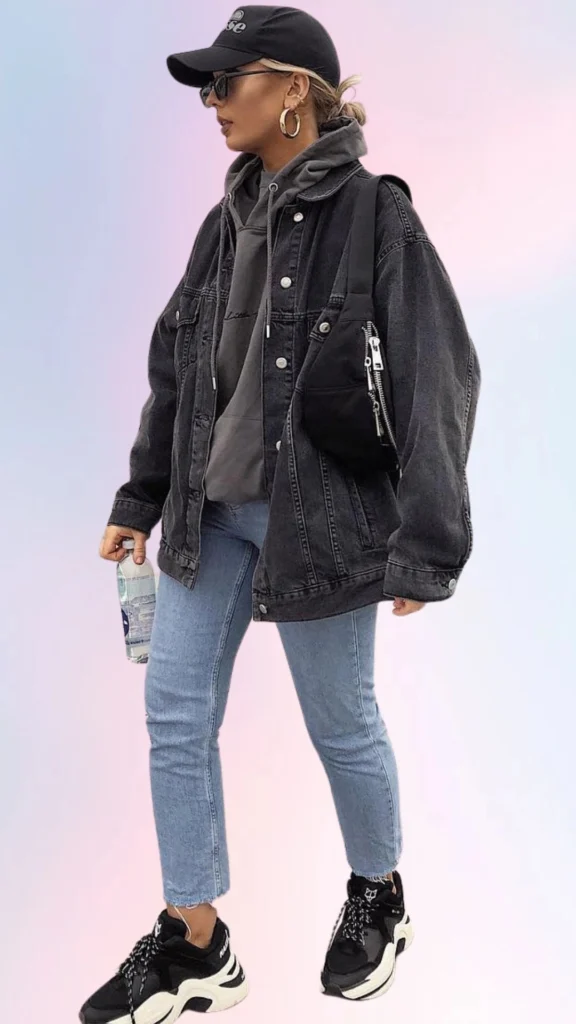 The outfit features a relaxed black denim jacket layered over a cozy gray hoodie, paired with light blue skinny jeans for a laid-back vibe. Completing the look, chunky black and white sneakers add a contemporary touch, while a black baseball cap and hoop earrings enhance the casual aesthetic. This ensemble is perfect for easy, comfortable days while still looking stylish.