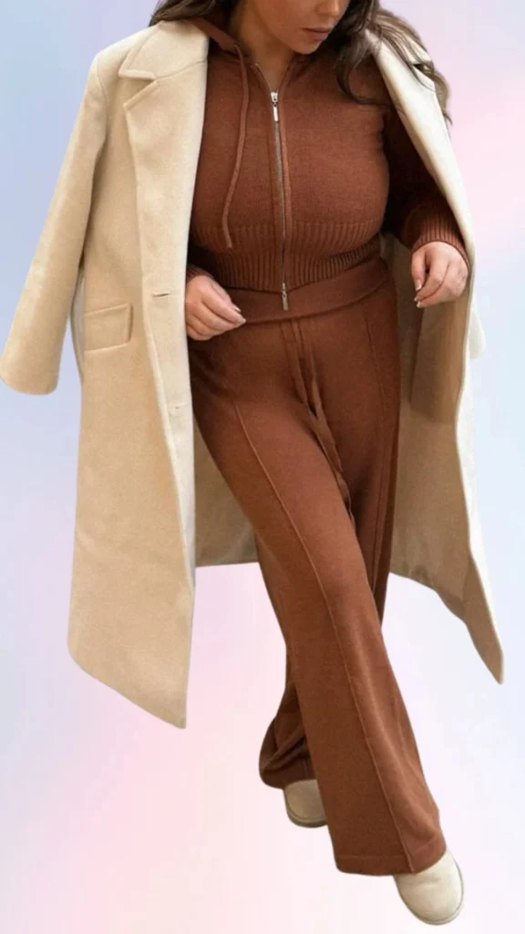 The outfit features a cozy, ribbed brown zip-up hoodie paired with matching wide-leg pants, creating a relaxed silhouette. A soft beige oversized coat drapes over the ensemble, adding warmth and a touch of sophistication. The look is completed with light cream slip-on shoes, embodying a laid-back yet stylish vibe perfect for easygoing days.