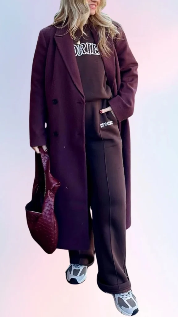 A relaxed outfit features a cozy brown sweatshirt layered under a long, rich burgundy overcoat, paired with matching dark joggers that have a subtle logo detail. The ensemble is complemented by comfortable gray sneakers and a slouchy burgundy tote bag, creating a laid-back yet stylish look. The soft textures and monochromatic color scheme enhance the chill vibe, making it perfect for a lazy day.