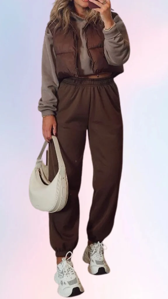 The outfit features a cozy gray oversized hoodie layered under a cropped, puffy brown vest, paired with relaxed, tapered brown joggers, creating a warm and inviting look. Complements include chunky white sneakers and a soft, cream-colored shoulder bag, enhancing the laid-back vibe. The textures of the fabrics and earthy color palette emphasize comfort while maintaining a stylish appearance.