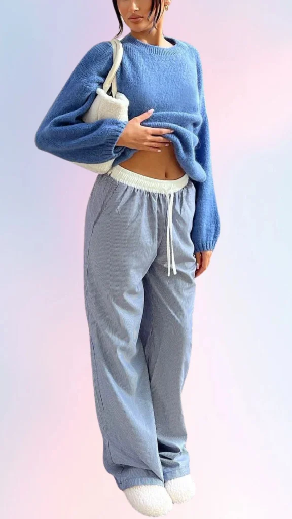 A cozy light blue oversized sweater features relaxed, voluminous sleeves and a soft texture, paired with striped, loose-fitting trousers that have an elastic waistband for easy comfort. The outfit is completed with fluffy white slip-on shoes, creating a laid-back and effortlessly chic look perfect for a lazy day. A small, textured bag adds a practical touch without sacrificing style.