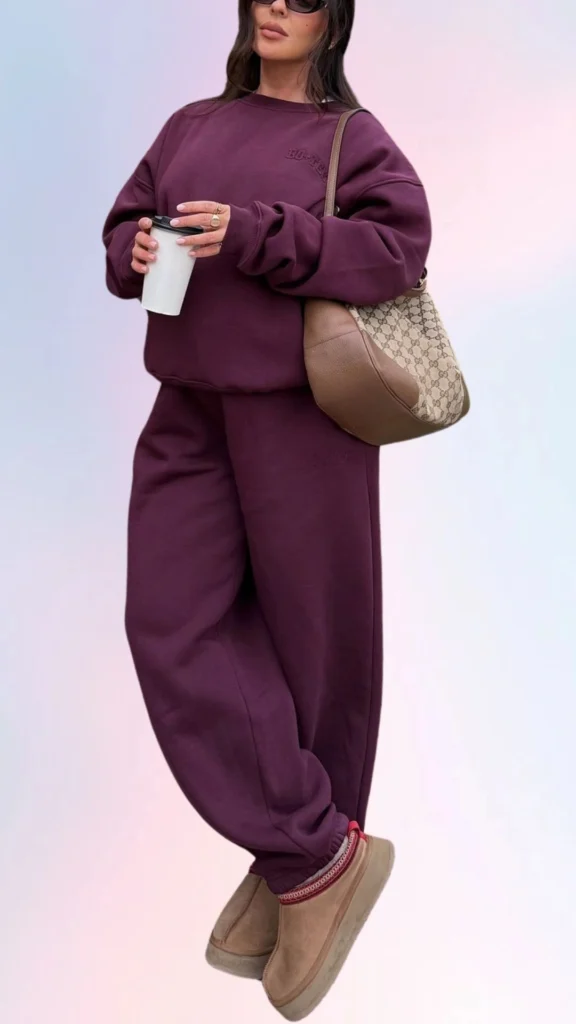 The outfit features a cozy, oversize maroon sweatshirt with subtle branding, paired with matching relaxed-fit sweatpants that emphasize comfort. Completing the look, soft beige slip-on shoes add a touch of warmth, while the oversized handbag provides practicality and style, ideal for a laid-back day. A warm beverage in hand enhances the relaxed vibe of this chic ensemble.