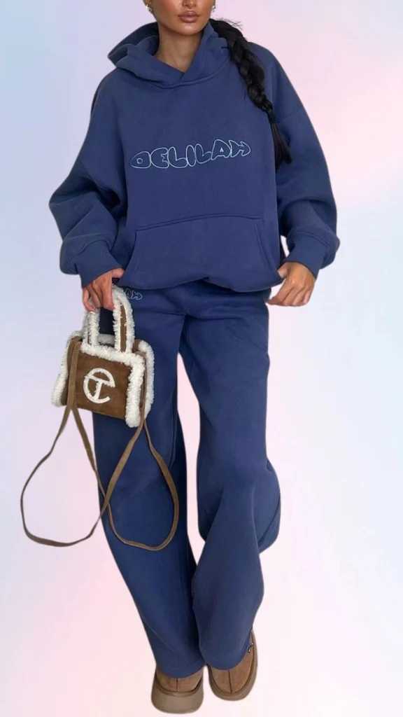 A cozy, oversized navy hoodie features a relaxed fit and playful logo, paired with matching baggy sweatpants for ultimate comfort. The outfit is complemented by soft, brown slip-on shoes and a compact, fuzzy handbag, adding a touch of casual chic to the laid-back ensemble. This stylish yet comfortable attire is perfect for a leisurely day at home or a casual outing.