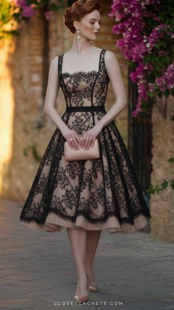 The image features a woman in a fitted, sleeveless cocktail dress with a nude base adorned with intricate black lace. She accessorizes with a soft pink clutch and wears elegant nude heels, complemented by dangling earrings, while standing in front of vibrant purple flowers in a sunlit outdoor setting. The dress's full skirt flares gracefully, enhancing its retro-inspired silhouette for a sophisticated look.