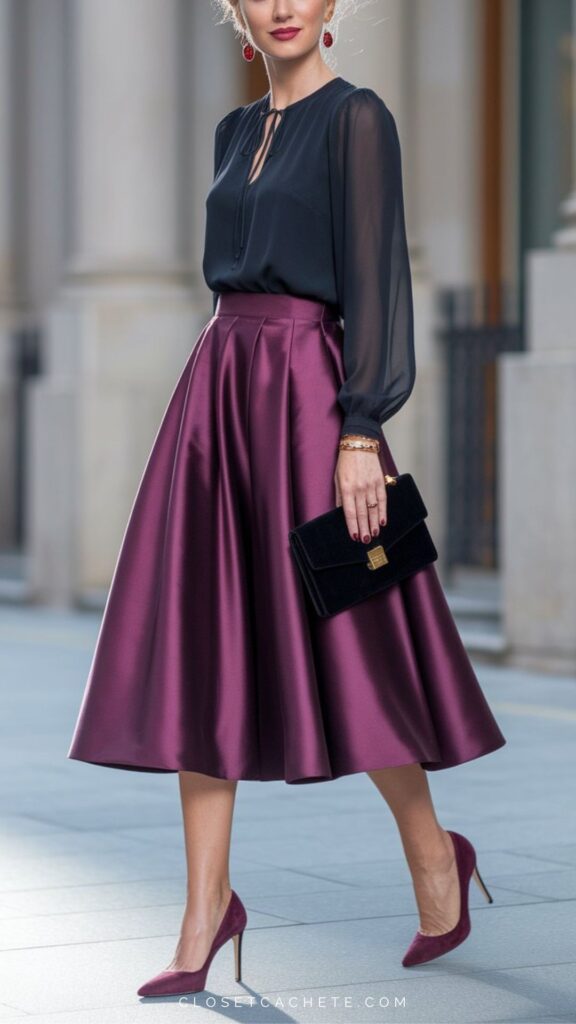 A woman is wearing a fitted black blouse with a tie closure at the neckline, paired with an elegant, full knee-length skirt in rich burgundy satin. She accessorizes with a small black clutch and matching burgundy high heels, complemented by bold red drop earrings, creating a chic cocktail attire look.