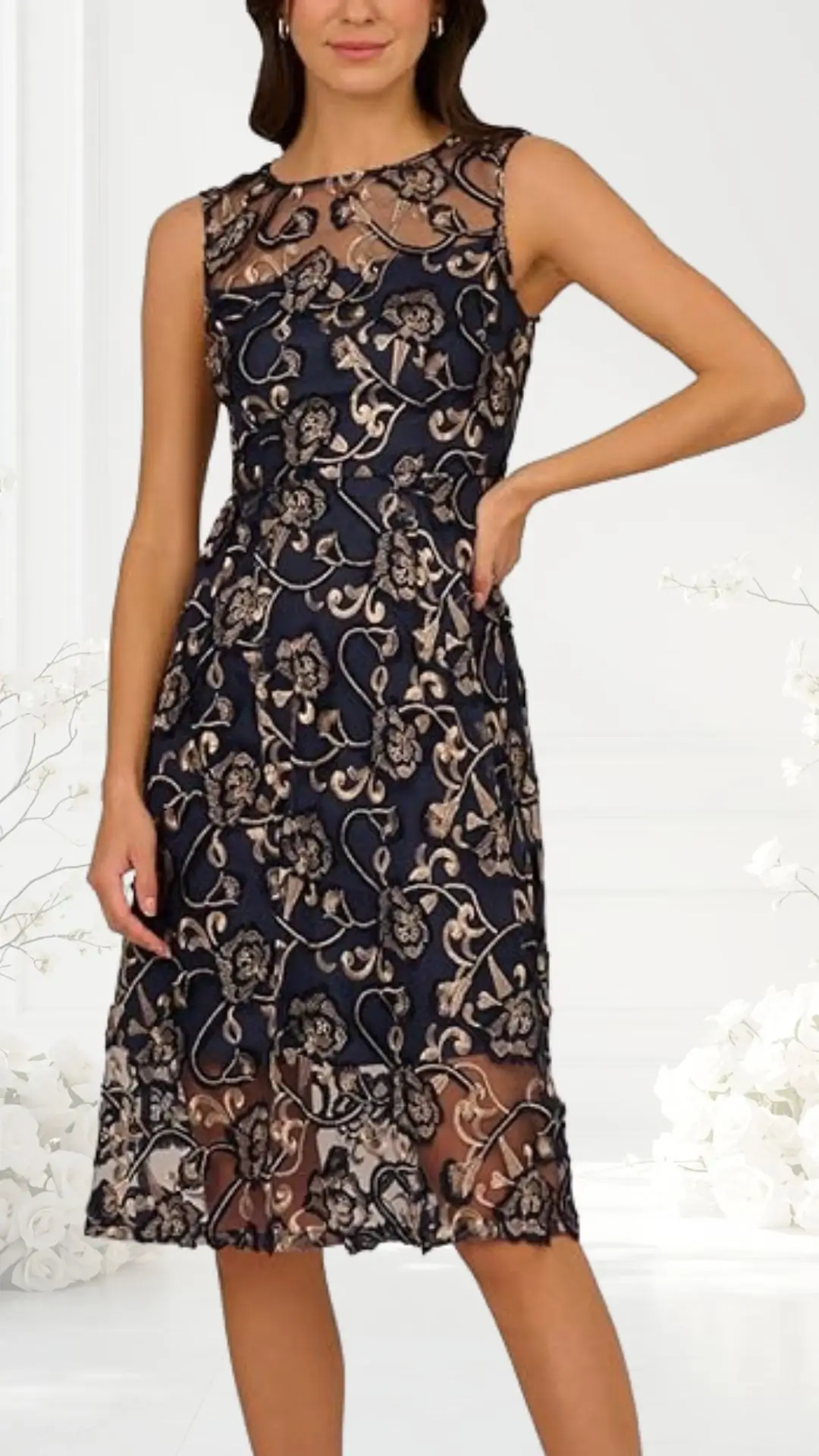 A sleeveless cocktail dress features a fitted bodice adorned with intricate floral embroidery in metallic gold and soft pink against a deep navy background. The skirt has a flowing silhouette that ends just below the knee, with a sheer overlay adding elegance. The model stands confidently with one hand on her hip, showcasing the dress's delicate design.