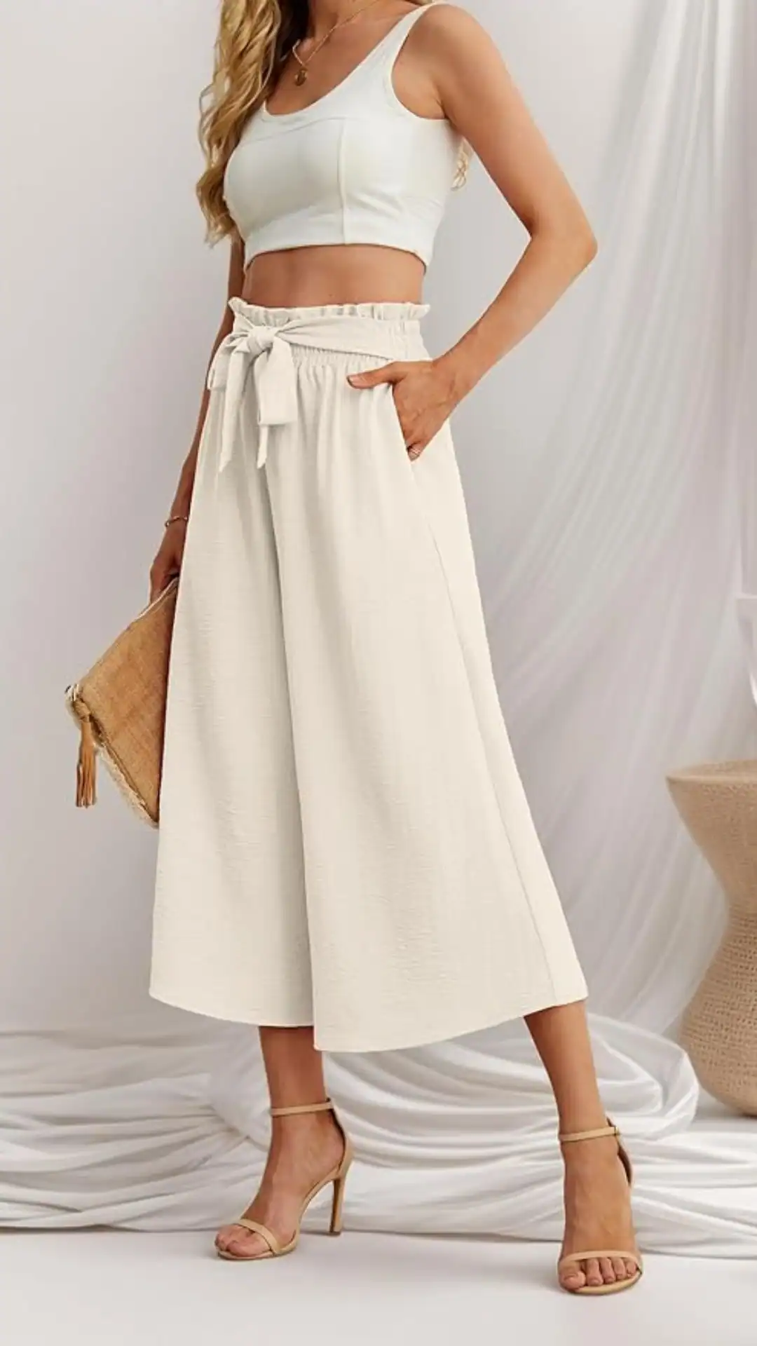 The outfit features a cream-colored linen matching set, comprising wide-leg trousers with a tie waist and a fitted crop top, creating a relaxed yet chic aesthetic. Its lightweight, textured fabric and neutral tone evoke a minimalist, summer vibe, suitable for casual outings or beach gatherings.