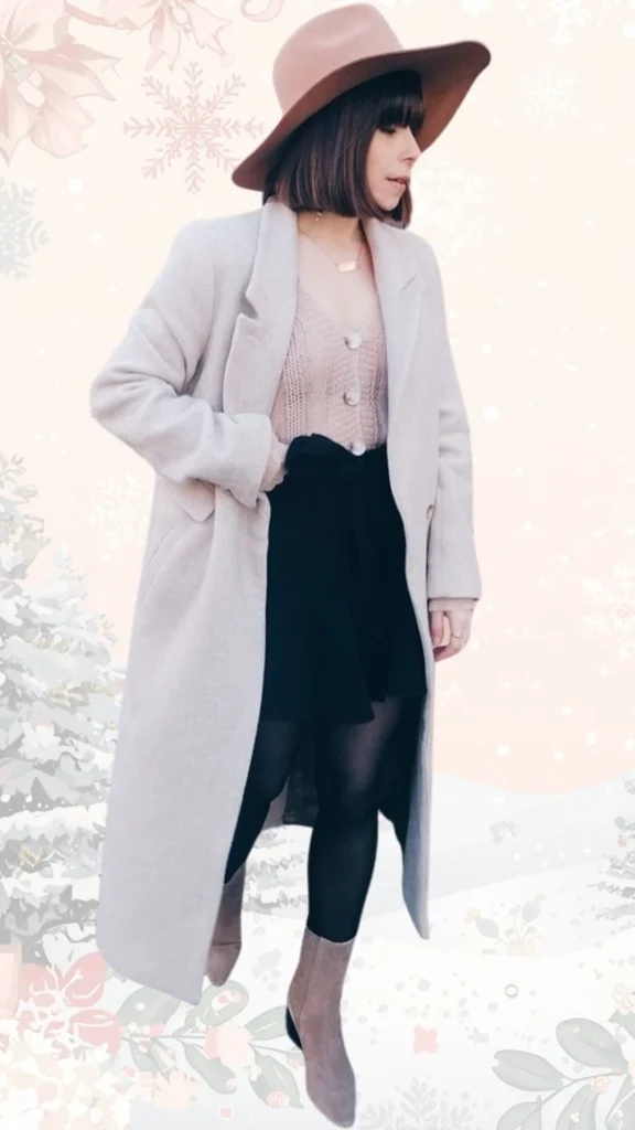 A light gray long coat is layered over a soft pink knit sweater adorned with buttons, paired with a black A-line skirt. She wears sleek black tights and stylish beige ankle boots, complemented by a wide-brimmed blush hat and delicate gold jewelry for an elegant winter look. The textures vary from the cozy knit to the smooth, polished appearance of the coat and shoes.