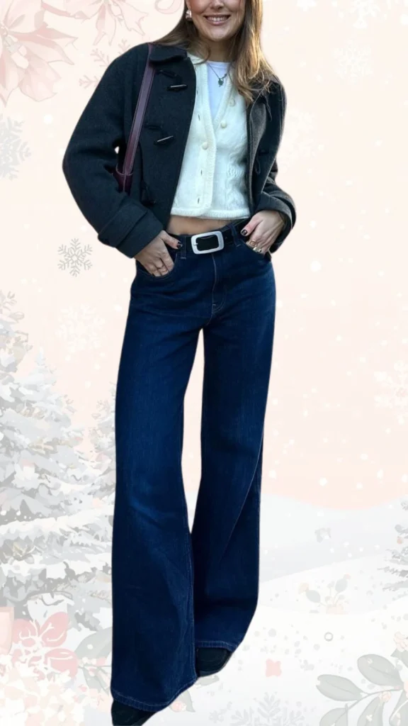 The outfit features a cozy, cream-colored cropped cardigan with a textured knit pattern layered over a simple white top, complemented by high-waisted, flared dark denim jeans. A stylish charcoal gray coat adds warmth, while a sleek black belt cinches the waist, and ankle boots provide a polished finish to the look. Accessories include silver rings and a chic shoulder bag, completing this cute and put-together winter ensemble.