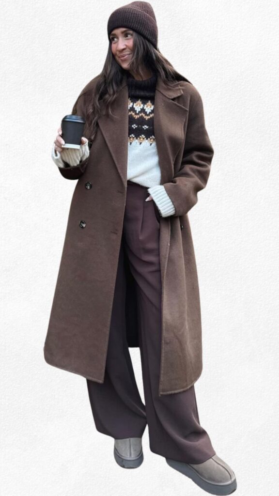 A woman stands outdoors in a stylish brown double-breasted coat over a cozy cream sweater featuring a festive geometric design in earthy tones. She pairs this with wide-leg maroon pants and gray plush ankle boots, accessorized with a matching brown beanie and holding a coffee cup, creating a warm and casual holiday-ready look. The scene is enhanced by subtle holiday decorations nearby.