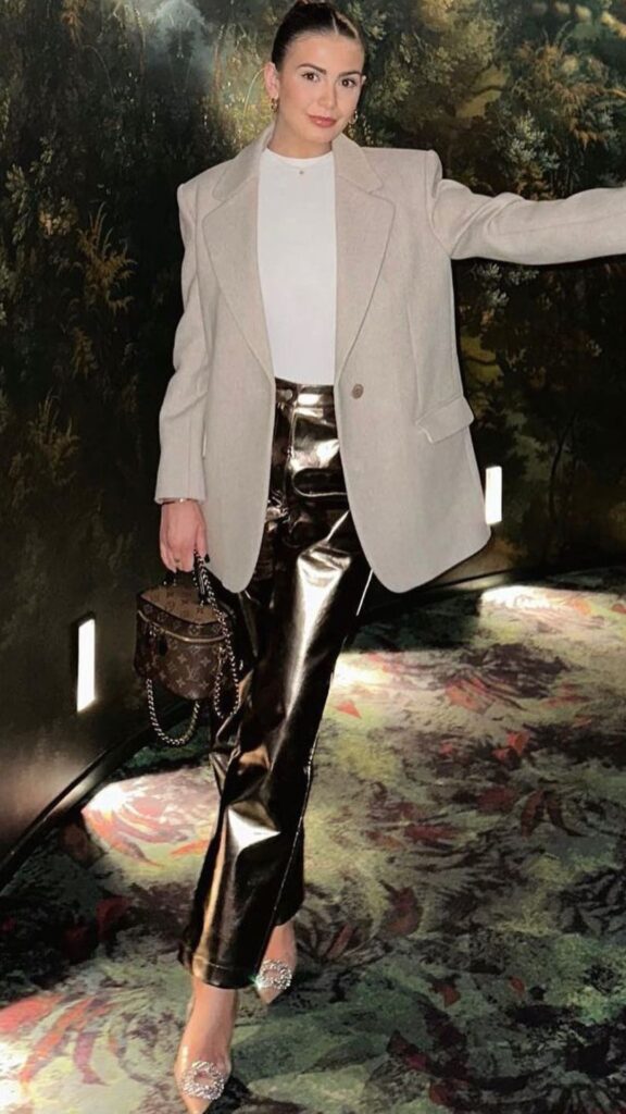 A tailored, oversized beige blazer adds sophistication, layered over a sleek white turtleneck for a crisp contrast. The standout metallic trousers feature a high-shine finish, creating a bold, modern silhouette, while elegant, translucent heels adorned with sparkling embellishments lend a chic touch, achieving a refined balance between contemporary style and timeless elegance.