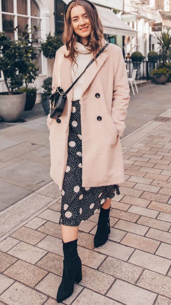 The outfit features a soft, blush pink double-breasted coat, paired with a cozy white turtleneck sweater and a flowy black midi skirt adorned with large white polka dots, creating a chic, feminine silhouette. Completing the look are sleek black sock boots that elongate the legs, adding a modern touch. The combination exudes a stylish yet timeless elegance, ideal for navigating the fall and winter seasons effortlessly.