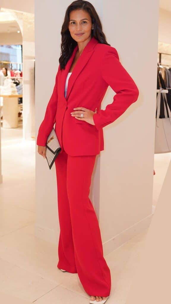 A structured, bold red suit comprising a tailored blazer with sharp lapels and matching wide-leg trousers exudes confidence and modernity. The ensemble is complemented by a minimalist white top and a chic black and white clutch, creating a striking yet sophisticated look ideal for fall and winter 2025/2026 trends, emphasizing a balance between vibrant colors and refined elegance. Footwear features sleek, open-toed heels that add a contemporary touch without detracting from the overall polished appearance.