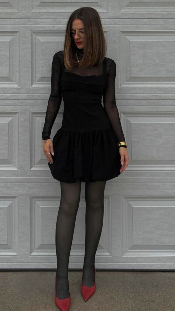 The outfit features a fitted long-sleeve black bodice with sheer lace sleeves, paired with a playful bubble skirt that adds volume and movement. It is completed with sheer black tights and striking red pointed-toe heels, creating a chic and timeless silhouette perfect for fall and winter 2025/2026. The overall look exudes sophistication and modern elegance while embracing the bubble skirt trend.