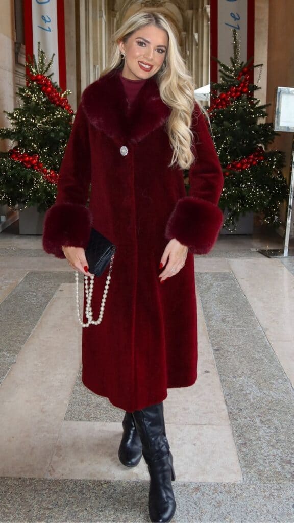 The outfit features a luxurious, long burgundy coat with plush fur cuffs and a sparkling brooch, seamlessly blending bold color with elegant details. Underneath, a fitted burgundy turtleneck complements the coat, while sleek black knee-high boots add a sharp contrast, enhancing the polished yet timeless aesthetic. The look is completed with a chic black handbag adorned with a string of pearls, embodying a sophisticated flair perfect for the fall and winter season.