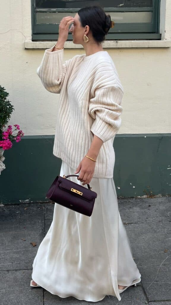 The outfit features an oversized ribbed cream sweater paired with a flowing, silky white maxi skirt that creates a sophisticated silhouette. A structured deep burgundy handbag adds a pop of color and serves as a statement accessory, while delicate gold hoop earrings complete the elegant yet relaxed look, making it a perfect blend of modern style with timeless appeal for the fall and winter seasons.