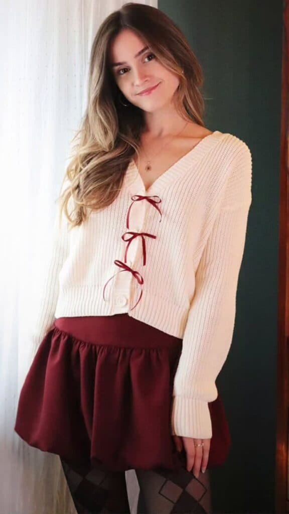 A cozy, textured cream cardigan with a V-neck and red ribbon detailing frames the look, paired with a rich burgundy bubble skirt that adds playfulness and femininity. Complete with patterned black tights, this outfit strikes a perfect balance between trendy and timeless, ideal for the fall and winter seasons without veering into costume-like territory. The overall vibe is effortlessly chic, showcasing soft color contrasts and layering for warmth and style.
