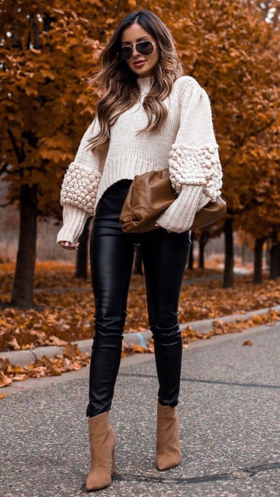 This outfit features an oversized cream knit sweater with textured puff sleeves, exuding a cozy yet sophisticated vibe. Paired with sleek black faux leather leggings that create a streamlined silhouette, the ensemble is complemented by tan ankle boots and a slouchy brown clutch, offering a contemporary touch that balances comfort and chicness for the fall and winter seasons. The overall look combines warmth and style, making it ideal for staying in fashion while ensuring timeless elegance.